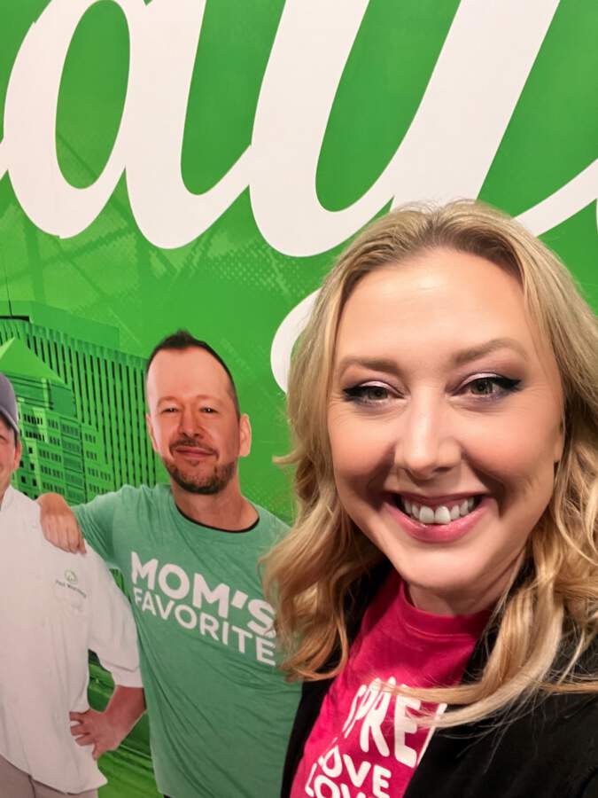 Writer, Nikki Nett, posing in front of green selfie station and mural of Mark, Paul, and Donnie Wahlberg at the Dayton Ohio location of Wahlburgers inside Hollywood Gaming and Racino  